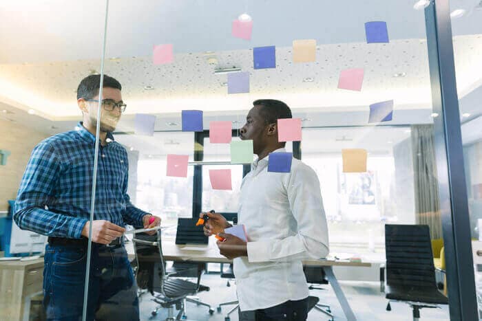 Two men talking in a modern startup office using a glass wall covered with sticky notes